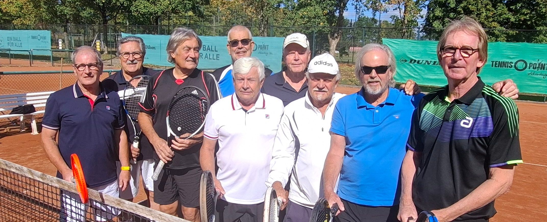 Neun Spieler der Herren 70 stehen mit ihren Schlägern am Netz auf einem Sandplatz, im Hintergrund Bäume und Banden.