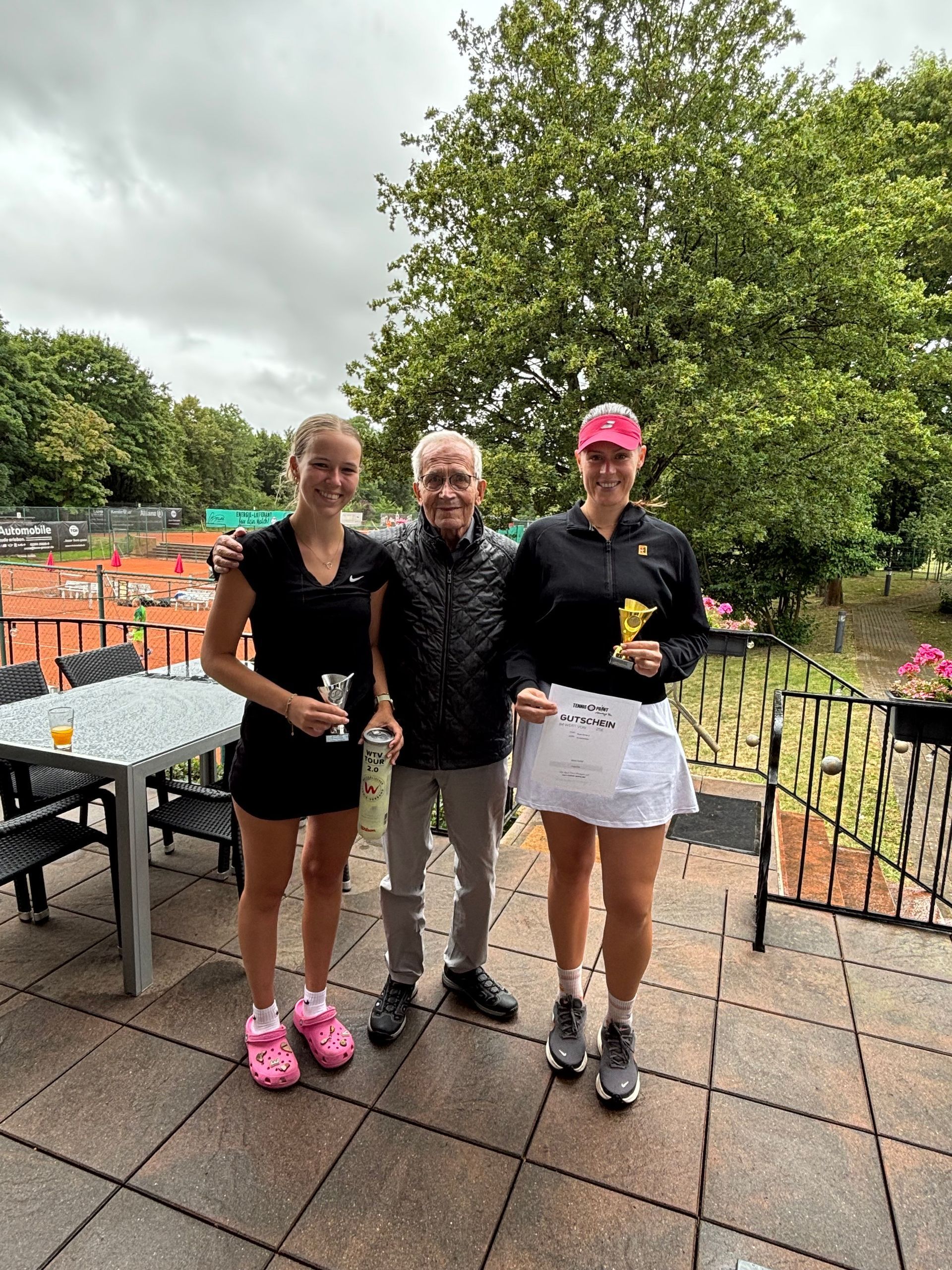 Zwei Tennisspielerinnen mit Pokalen und Turniernamensgeber Horst Zweydinger auf der Terrasse, im Hintergrund die Plätze.