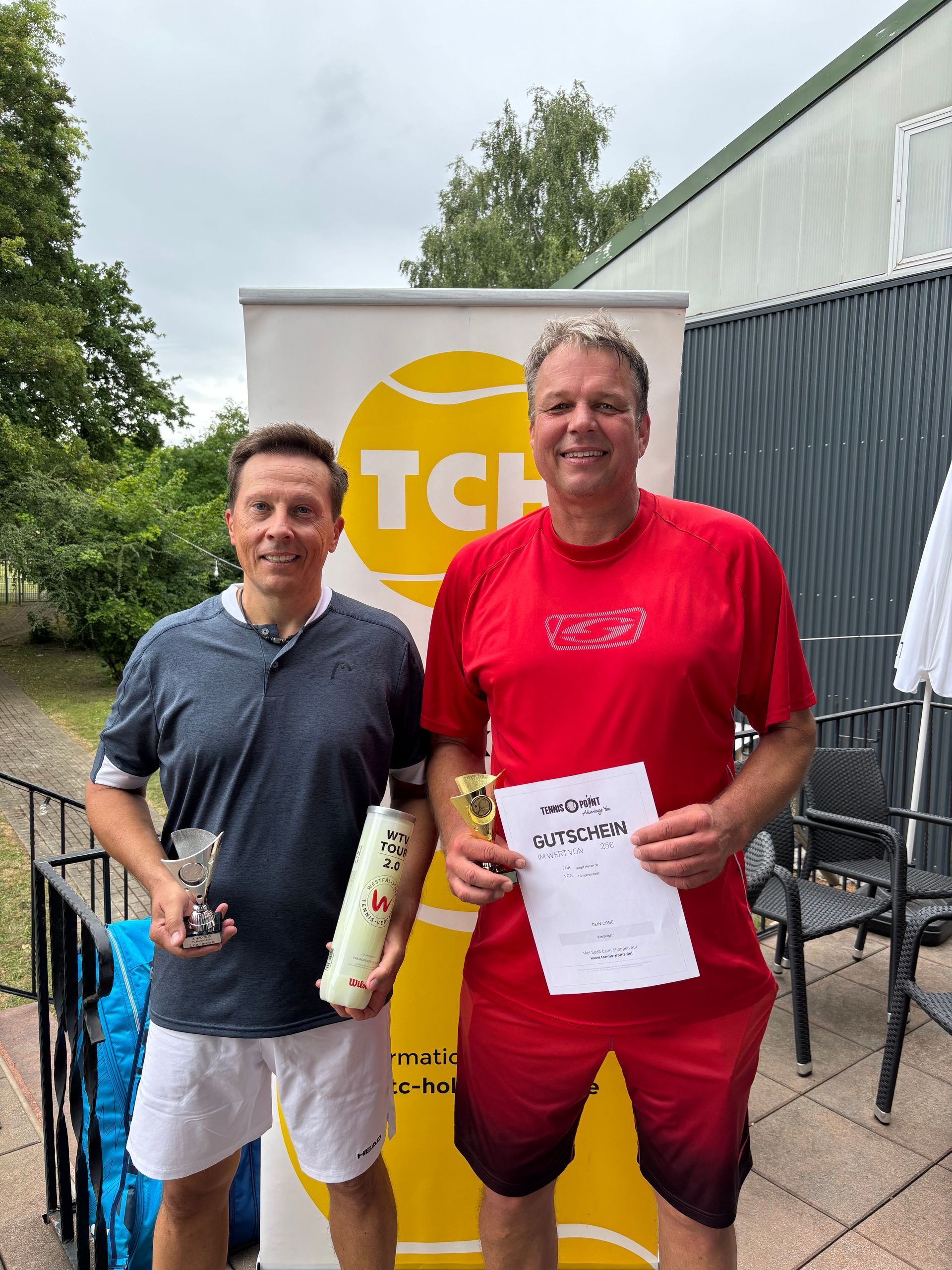 Zwei Tennisspieler mit Pokal und Gutschein vor einem TCH-Aufsteller auf der Terrasse.
