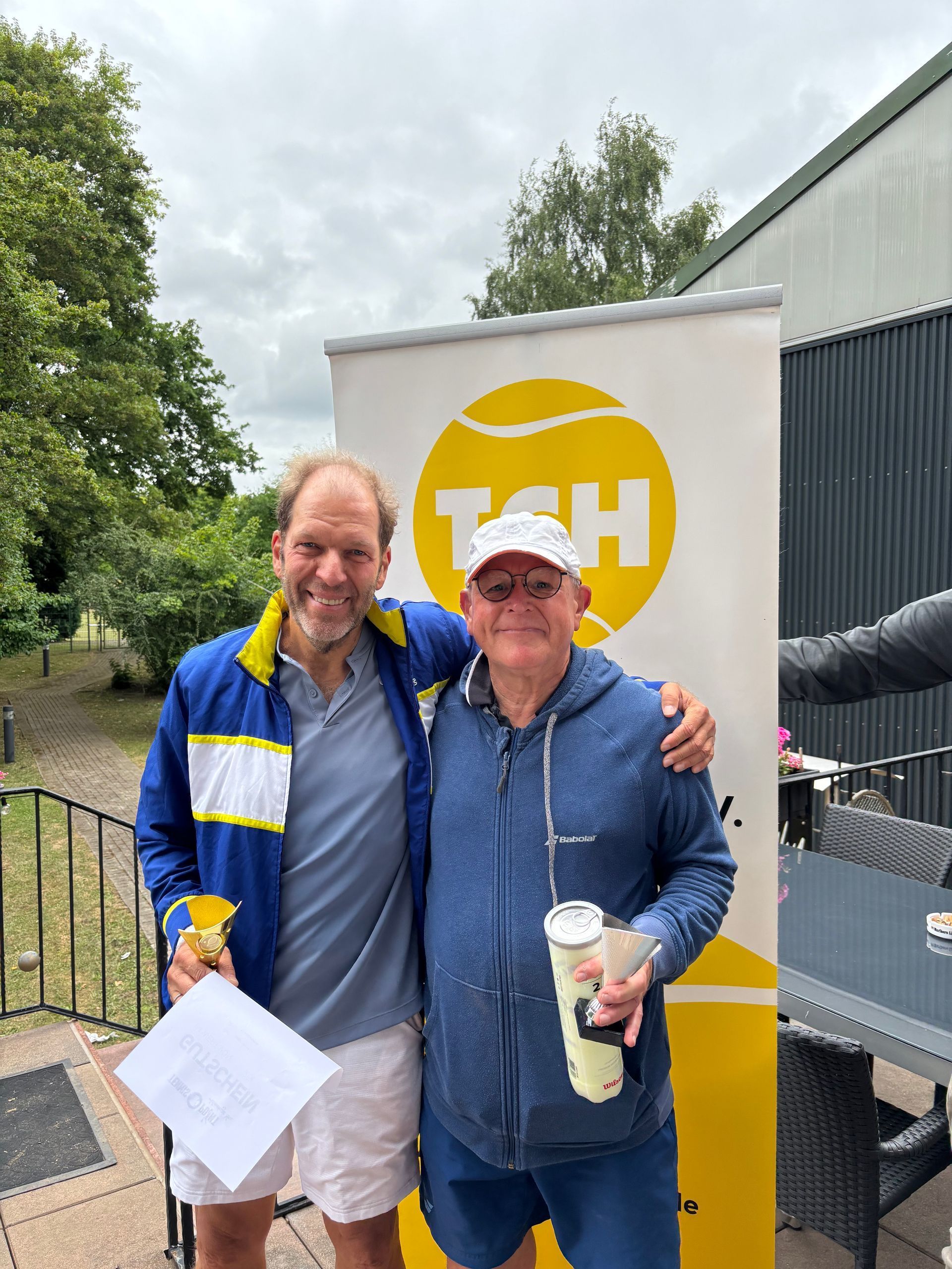 Zwei ältere Tennisspieler mit Urkunde und Pokal vor einem TCH-Aufsteller.