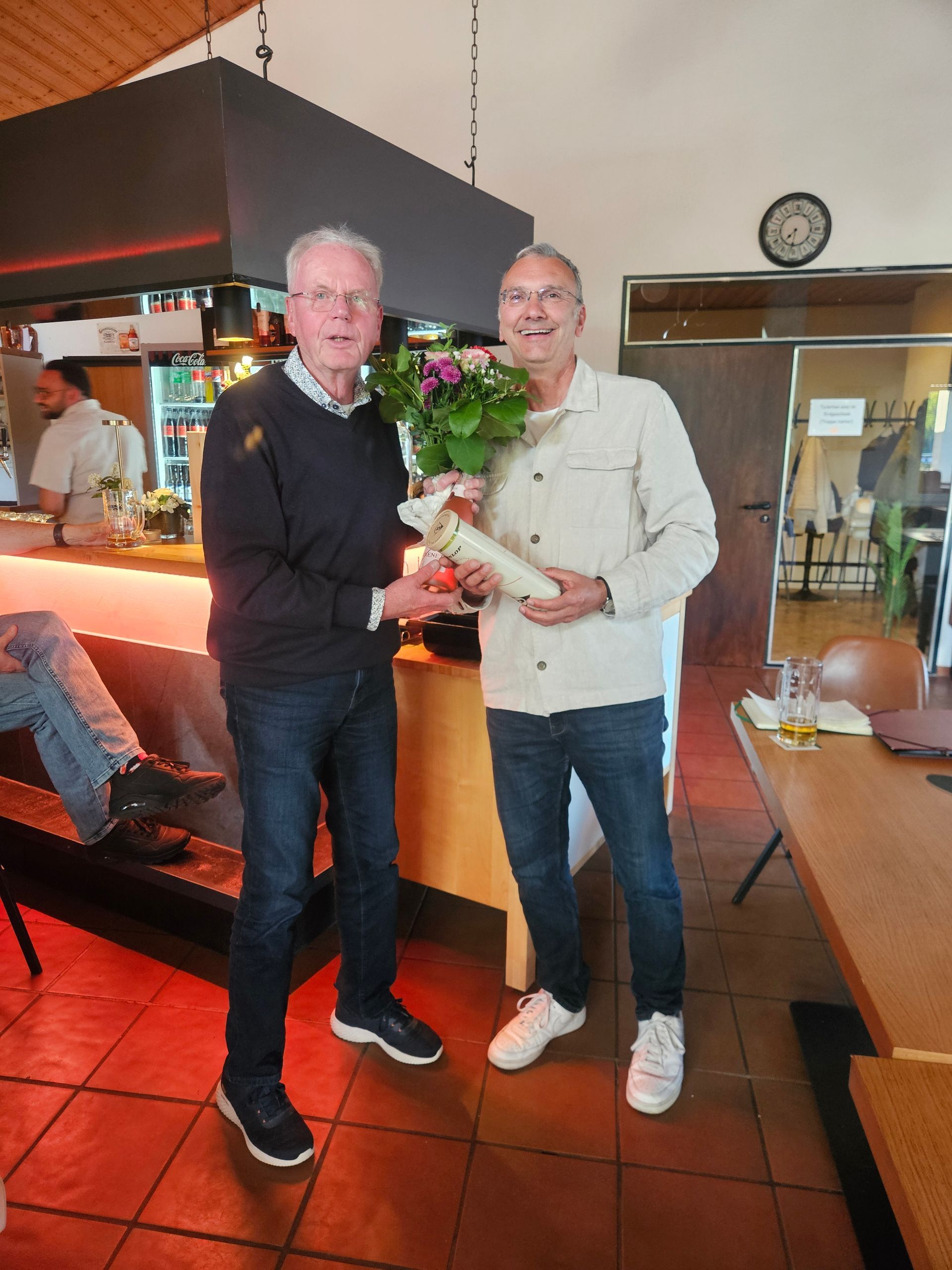 Zwei Männer im Clubrestaurant, einer hält einen Blumenstrauß.