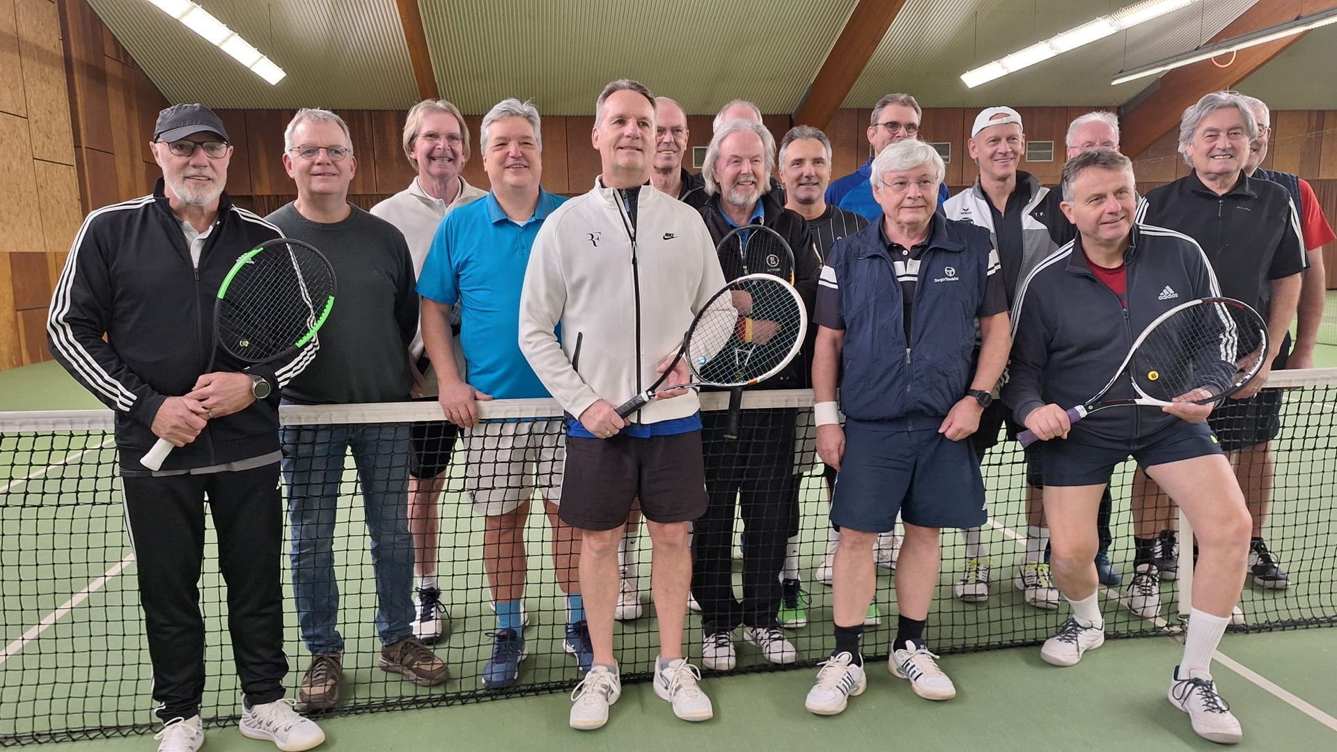 Gruppenfoto von 16 Tennisspielern mit Schlägern vor dem Netz in der Tennishalle.
