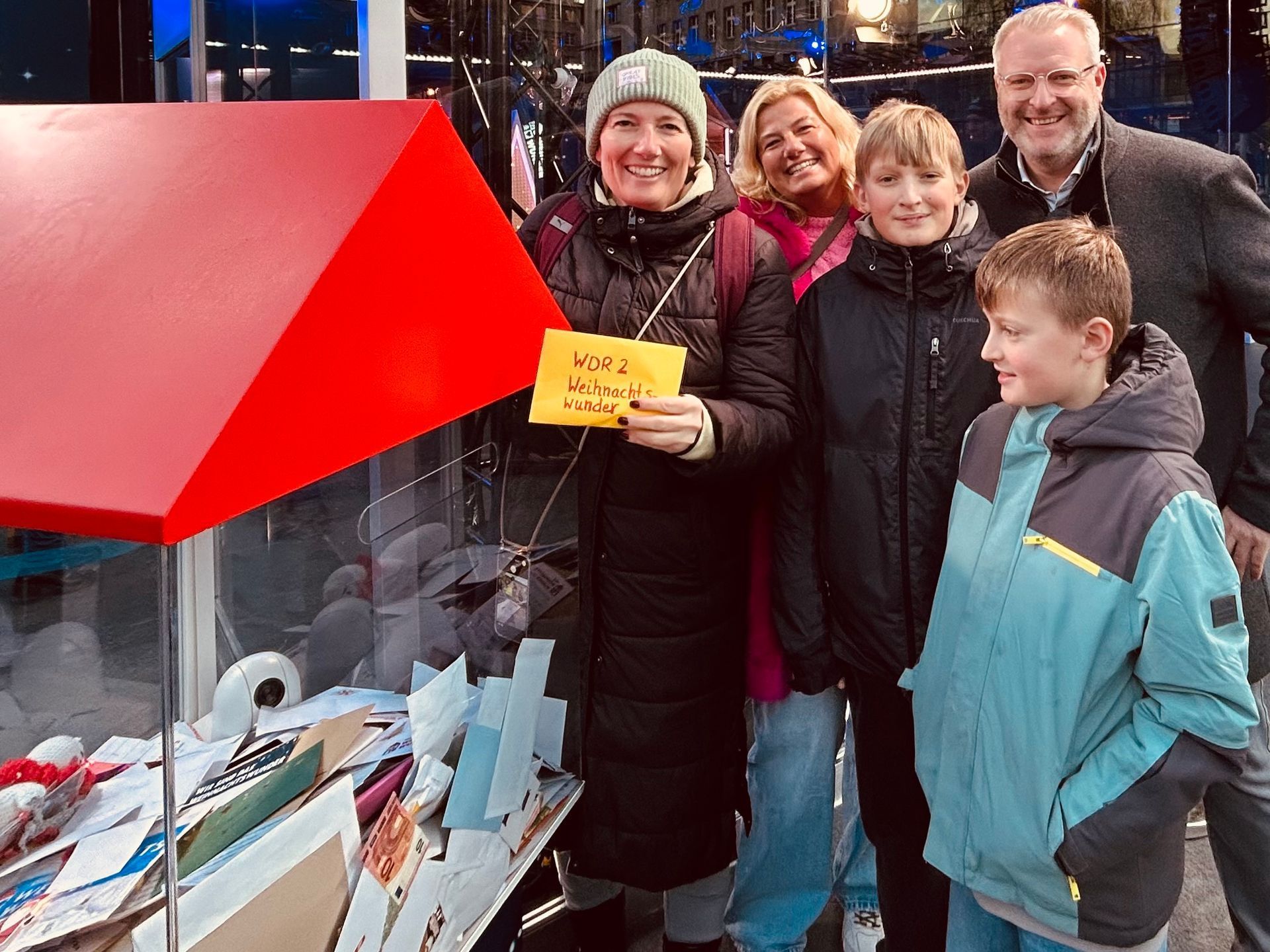 Kinder und Erwachsene stehen beim WDR 2 Weihnachtswunder vor einem roten Spendenhäuschen, eine Frau hält einen Umschlag.